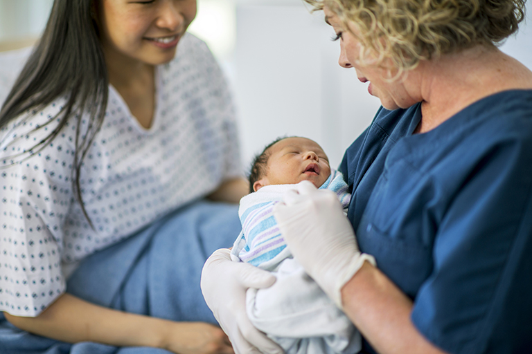 A nurse holding a sleeping newborn baby in her arms while the mother sits on the hospital bed