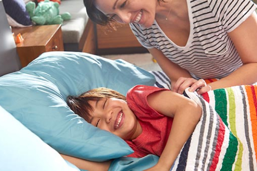 A smiling mother tugging her laughing child under a colorful blanket in bed.