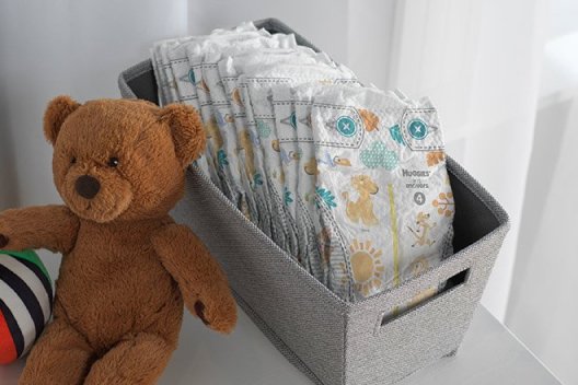 A gray tote full of Huggies Little Movers Diapers sits on a dresser next to a teddy bear and a plush ball