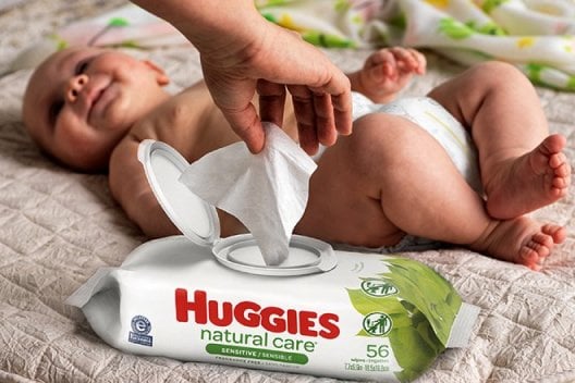 A smiling baby lays on a blanket while a hand pulls a wipe from a package of Huggies Natural Care Sensitive Wipes with EZ Pull lid
